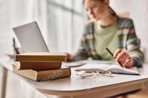 Preview: focus on books on desk, teenage girl holding pen and looking at laptop while studying from home