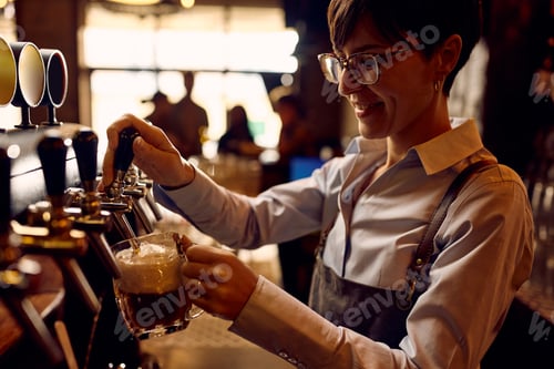 Preview: Happy female barista pouring beer from a beer tap while working in pub.