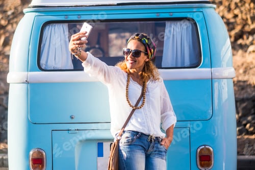 Preview: One young adult woman taking selfie picture with phone against a blue classic van. Travel