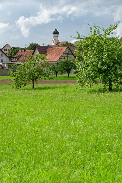 Preview: Vertical shot of an idyllic rural German village in the backdrop situated in a lush green meadow