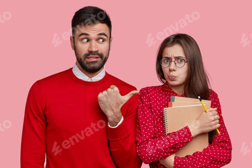 Preview: Handsome unshaven man wears red jumper, points at displeased beautiful woman who makes grimace, carr