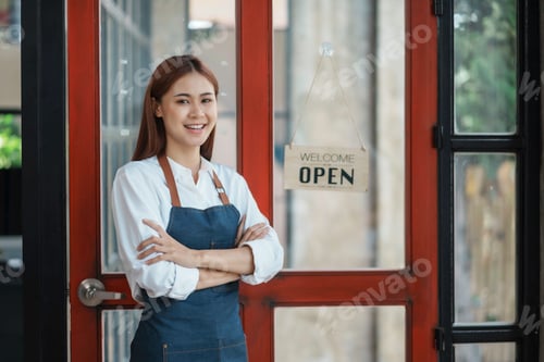 Preview: Charming Asian businesswoman in an apron waits to open a shop in front of a cafe with a beaming smil