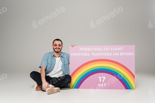 Preview: Cheerful gay man near placard with International Day Against Homophobia, Transphobia and Biphobia