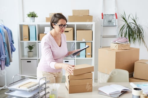 Preview: Young online shop manager with list of orders taking top box of stack