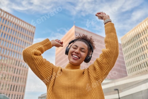Preview: Positive carefree curly haired woman dances with joy enjoys favorite audio track via headphones dres