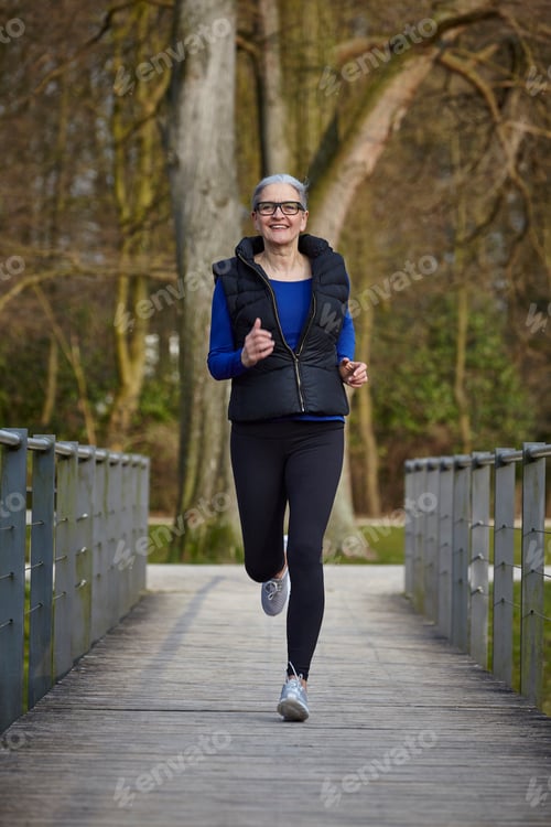 Preview: Full length front view of woman on wooden path jogging, looking at camera smiling