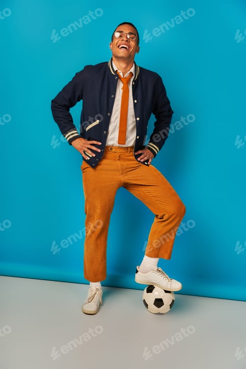 Preview: excited and trendy african american student stepping on soccer ball and laughing on blue
