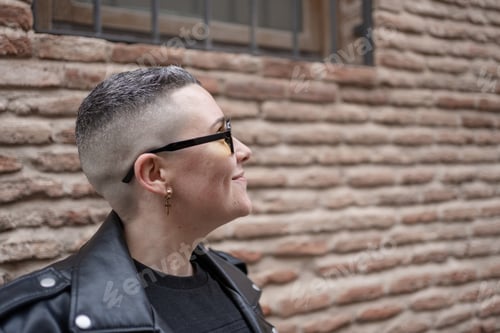 Preview: Smiling butch lesbian with shaved hair and black leather jacket posing near brick wall