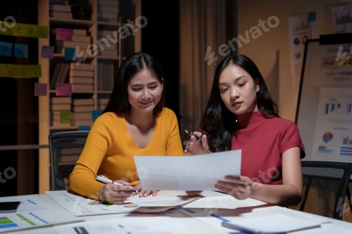 Preview: Two asian businesswomen working late in a dimly lit office, collaborating on a project