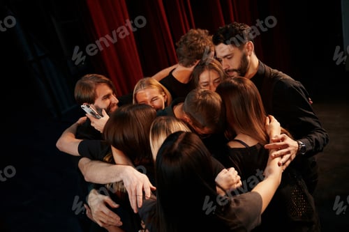 Preview: Embracing each other. Group of actors in dark colored clothes on rehearsal in the theater
