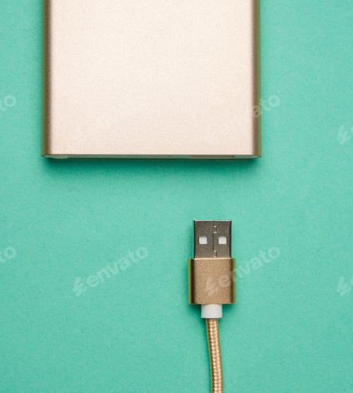 Preview: Golden power bank and cord with a usb connector for recharging mobile devices on a green background