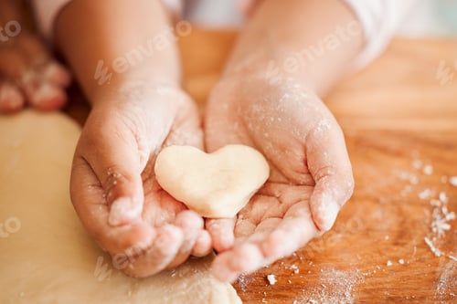 Preview: Closeup of an unknown chid holding a heart shaped cookie while baking in the kitchen with her mothe
