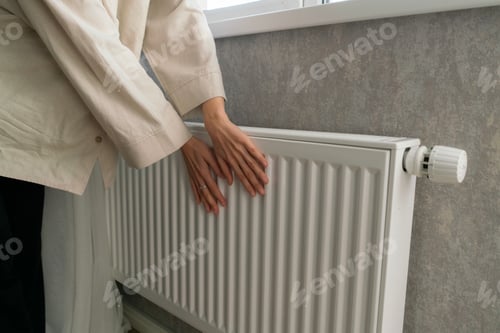 Preview: A woman huddles close to a radiator, seeking warmth