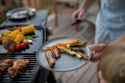 Preview: Father putting grilled meat and vegetable on plate to his son during family summer garden party.