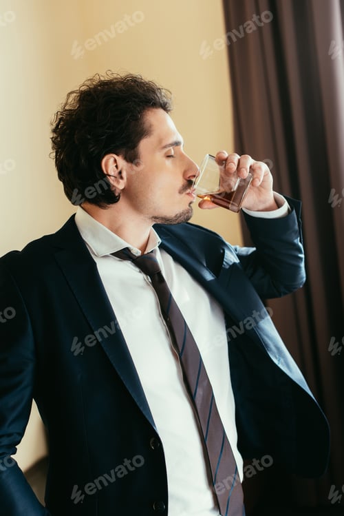 Preview: businessman in suit drinking whiskey from glass in hotel room