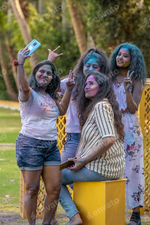 Preview: Group of young women celebrating Holi festival with colorful powder, taking a selfie in a park