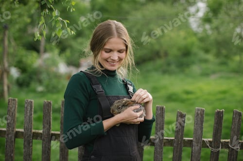 Preview: Young beautiful woman farmer with a baby rabbit in her hands on the background of the countryside.