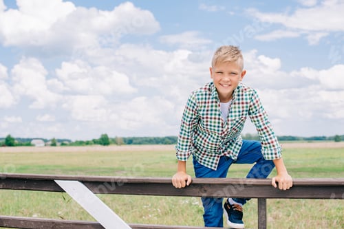 Preview: happy boy climbing on fence and smiling at camera on farm