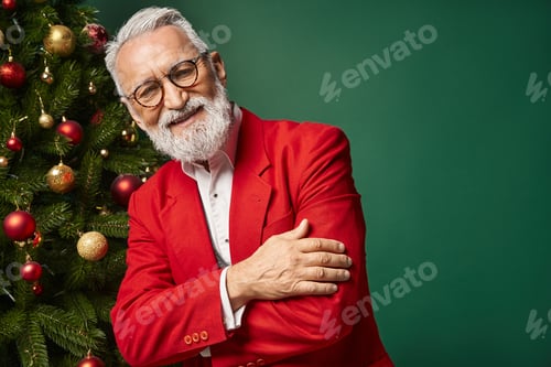 Preview: cheerful man in classy Santa suit smiling at camera next to Christmas tree, winter concept