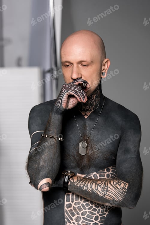Preview: bald shirtless man with tattoos smoking cigar and looking down on grey