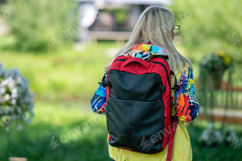 Preview: blonde middle-aged woman in bright clothes with a backpack in the park on a sunny day, rear view