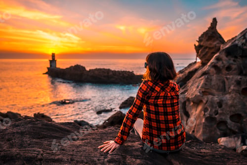 Preview: A young woman sitting on some rocks with a leather jacket in the orange sunset
