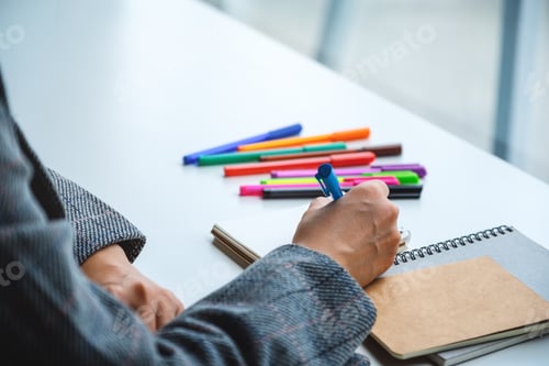 Preview: Closeup image of a woman writing on a blank notebook with colored pens on the table
