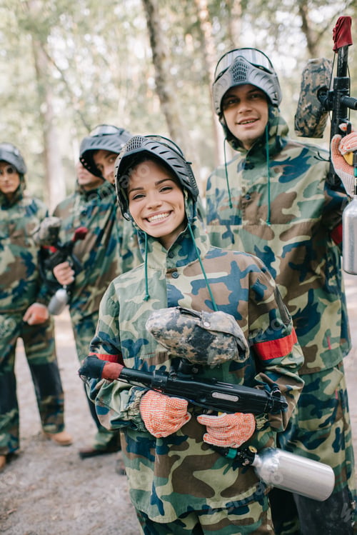 Preview: beautiful happy female paintballer in uniform holding paintball gun while her team standing behind