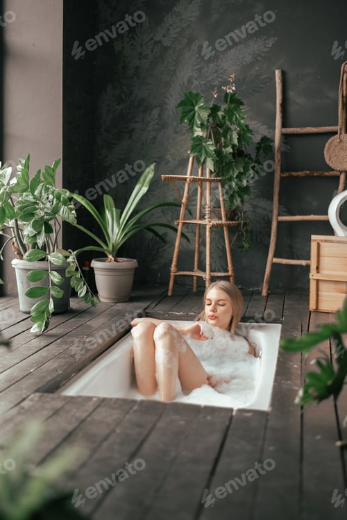 Preview: Young beautiful blonde woman taking bath with bubbles in the luxury bathroom decorated with plants