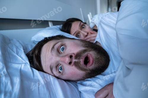 Preview: Man had terrible nightmare, woke up in horror, open mouth and eyes, frightened woman looks at him