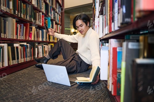 Preview: Relaxed Japanese man studying laptop, holding paper book and smartphone