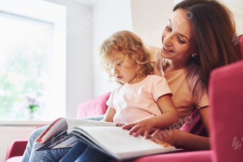 Preview: Reading big book. Young mother with her little daughter in casual clothes together indoors at home