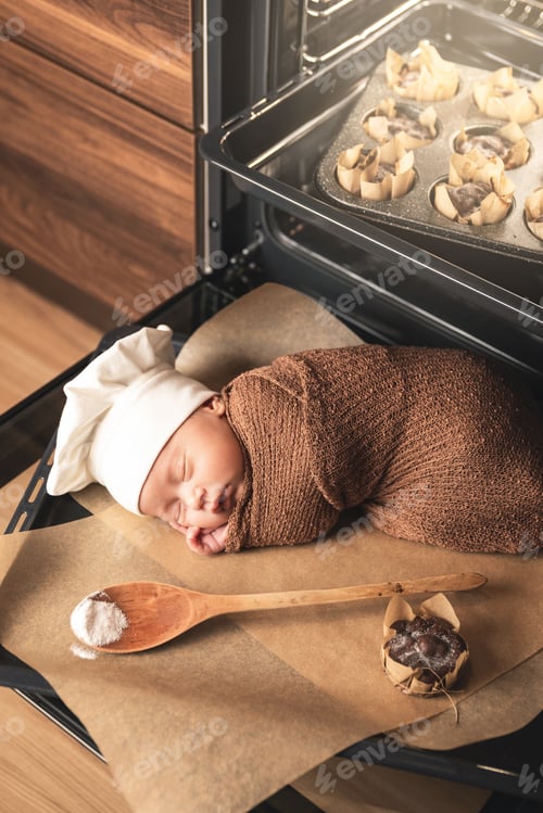 Preview: Newborn baby wearing chef's hat is lying on the oven tray with a muffins