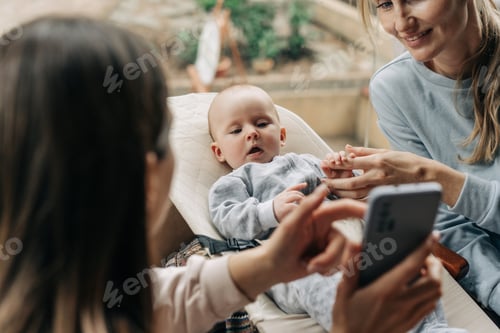 Preview: Women sitting in a cafe and nursing a baby take photos on their phones and look at them.