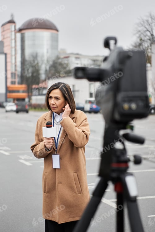 Preview: Multiracial journalist in beige coat reporting news outdoors in urban setting on a cloudy day
