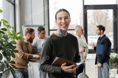 Preview: Cheerful tattooed businessman holding paper folder and looking at camera near interracial