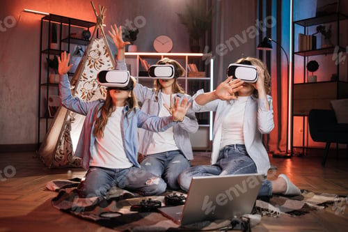 Preview: Happy mother and cute daughters sitting near toy wigwam at home and using modern VR headset