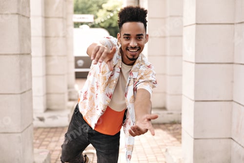 Preview: Stylish charming young curly dark-skinned man in black jeans and floral shirt points at camera and