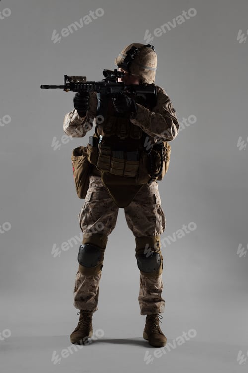 Preview: Military personnel stands ready with weapon in studio against gray background