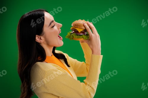 Preview: side view of young and hungry woman opening mouth near burger with cheese and meat isolated on green
