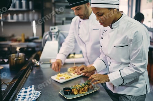 Preview: Black female chef and her colleague serving food while working at restaurant kitchen.