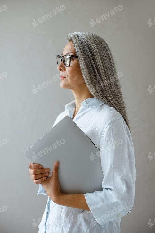 Preview: Serious mature Asian businesswoman holds laptop standing near grey stone wall