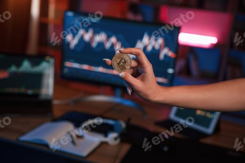 Preview: Close up view of woman's hand that holding bitcoin against computer display and tablet with graphs