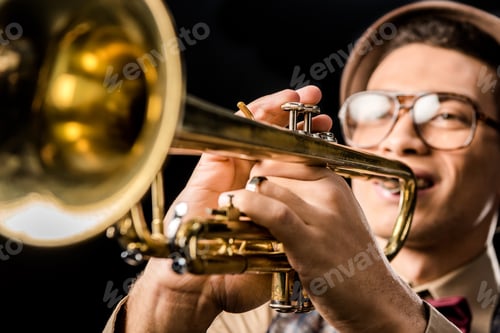Preview: selective focus of male jazzman in hat and eyeglasses playing on trumpet isolated on black