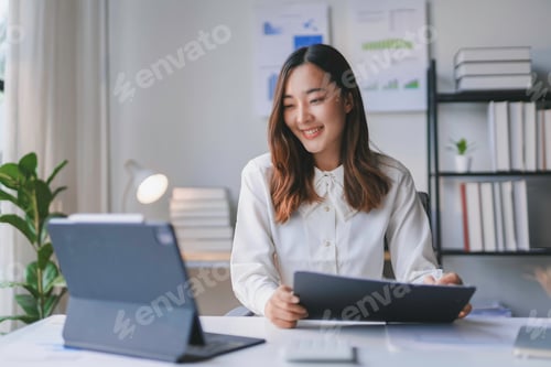 Preview: Asian businesswoman is smiling while working from her home office and using a tablet computer