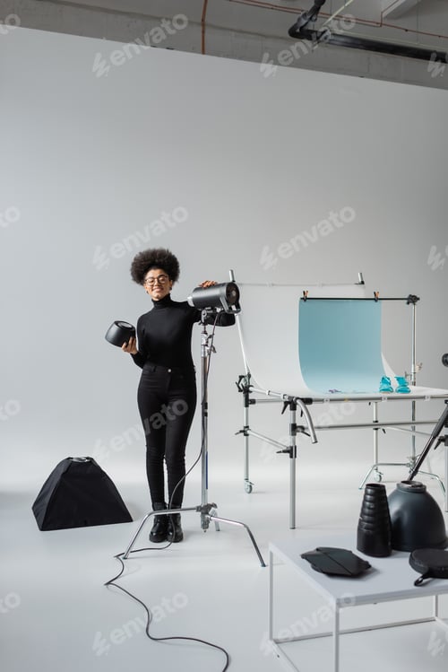 Preview: full length of happy african american content maker near strobe lamp and shooting table in