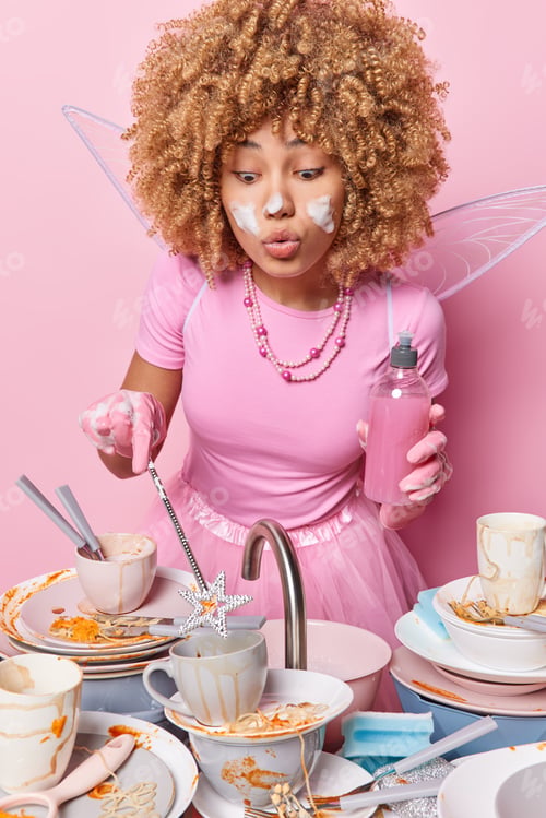 Preview: Surprised curly haired woman holds magic wand tries to make in such away dirty dishes become clean