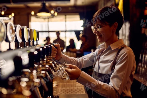 Preview: Happy female barista pouring lager beer from beer tap at bar counter.