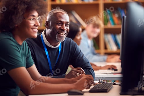 Preview: Male University Or College Student Working At Computer In Library Being Helped By Tutor