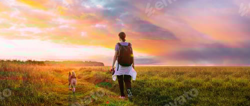 Preview: Young Woman Backpacker Walking With Dog In Summer Meadow Grass During Sunset Sunrise Time. Scenic
